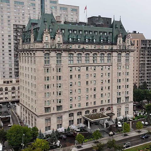 A photo of a grand, multi-story hotel with a light-colored stone facade and a distinctive green steeple roof, surrounded by modern high-rise buildings in an urban setting. The hotel features arched windows and a prominent entrance with a canopy, with trees and vehicles visible on the street below, and a Canadian flag atop the building.