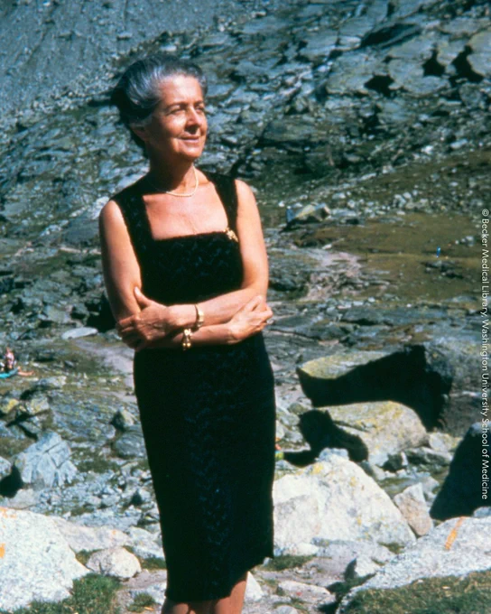 Rita Levi-Montalcini standing outdoors, smiling in a sunny setting with rocky terrain in the background.