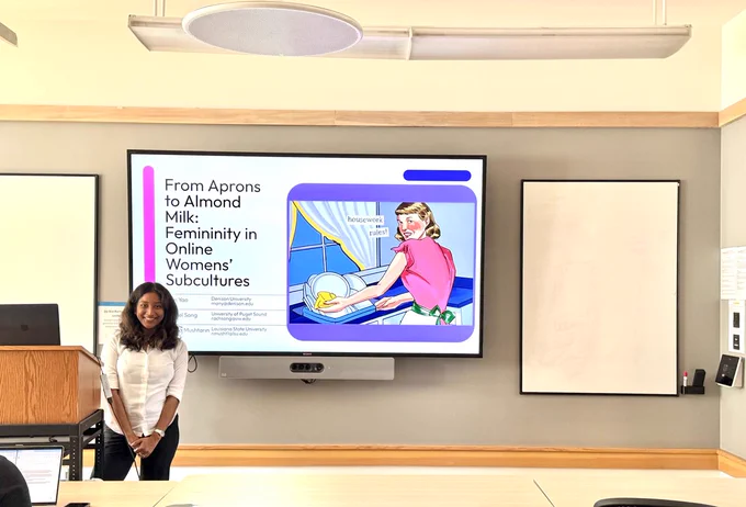 Nabila in front of the title slide to her research group's powerpoint presentation, "From Aprons to Almond Milk: Femininity in Online Womens' Subcultures"