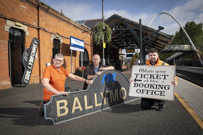 3 men on the train platform at Ballymoney, two are propping up an old Ballymoney station name sign. The third man is holding a sign saying "Obtain your ticket at the ticket office."