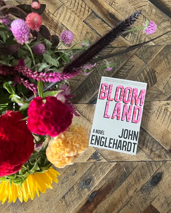 A paperback copy of BLOOMLAND by John Englehardt sits on a wooden table next to a bouquet of flowers. 