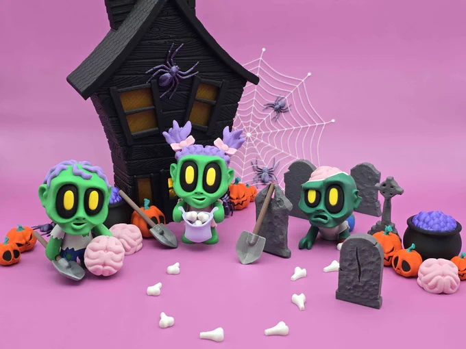 Three 3D-printed Blob Lab zombies posed in front of a haunted house with tombstones, pumpkins, spiderwebs, and bones on a pink background.