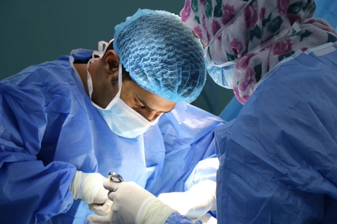 Two surgeons in blue scrubs and gloves performing a surgical procedure under bright operating room lights.