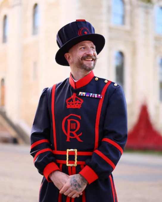 A Yeoman Warder in uniform smiling in front of the White Tower at the Tower of London.