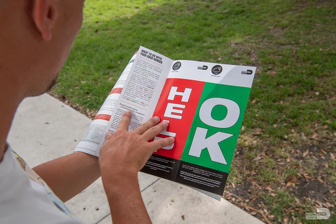 A person is reading a brochure titled "WHAT TO DO WITH YOUR DOOR HANGER." The brochure features large letters spelling "HELP" in red and "OK" in green, advising residents how to signal for assistance after a storm.