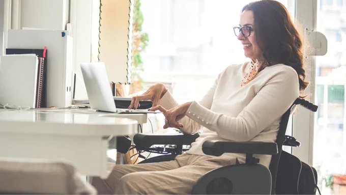 A woman in a wheelchair is smiling and using a laptop at a desk. She wears glasses, a light-colored sweater, and beige pants. Natural light fills the room from a large window.