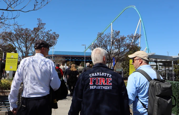 Three people walking towards the entrance of an amusement park, with one person wearing a jacket labeled "Charlotte Fire Department." A large roller coaster is visible in the background under a clear blue sky.