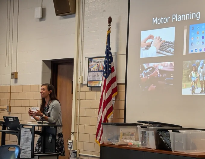 A woman is standing towards the front of a conference room. She is smiling. A projected screen with the title "Motor Planning" is behind her.