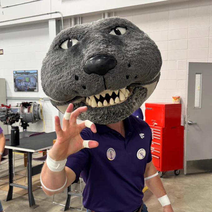 The K-State mascot, Willie the Wildcat, holds up the "WC" Wildcat hand gesture inside of the K-State Salina aviation maintenance hangar. 