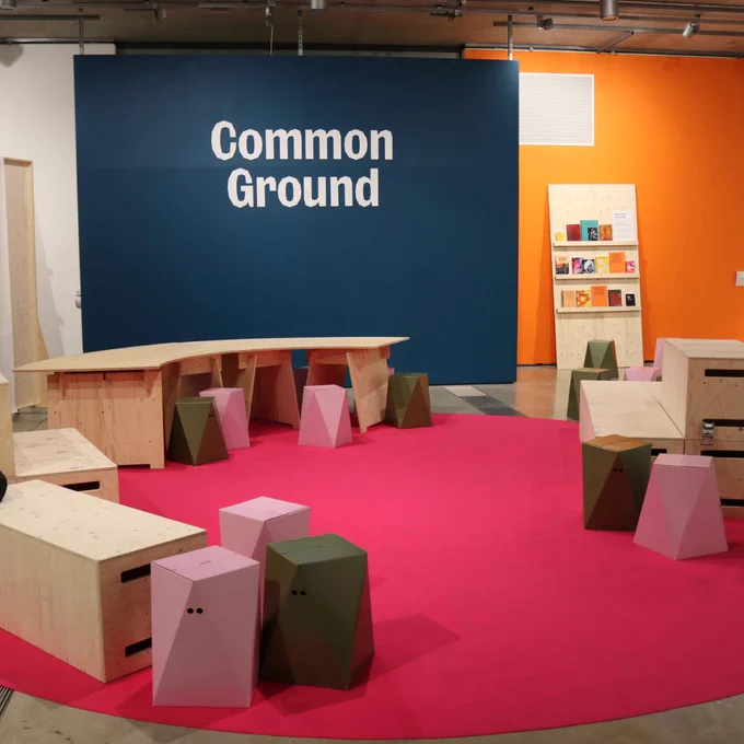 Exhibition space with wooden seating and tables on a bright pink floor, titled Common Ground, at Lethaby Gallery.