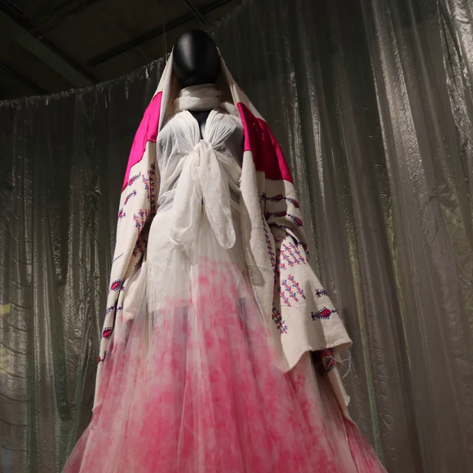 Mannequin wearing a layered white and pink dress with an embroidered shawl, displayed against silver curtains.