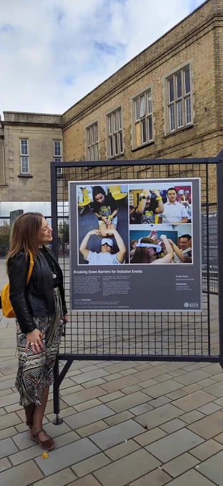 A woman (me) stands outside Bath Spa train station beside a large display board featured in the University of Bath’s Images of Research 2025 exhibition. The board showcases a collaborative project with Karin and Michael focused on identifying barriers and enablers when organizing accessible events for youth with vision impairment. The poster includes a collage of three classroom photos showing students with partial or total vision loss actively engaging with virtual reality headsets. In the left photo, a male student lies on the floor wearing a VR headset while a female student observes nearby. In the top right, the same female student wears the headset, raising her hands to interact with the virtual environment. In the bottom right, a group of students—including a blonde girl and a boy with ginger hair—wear VR headsets and raise their hands, with an adult smiling faintly in the background. The classroom has teal walls and an exit sign is visible.