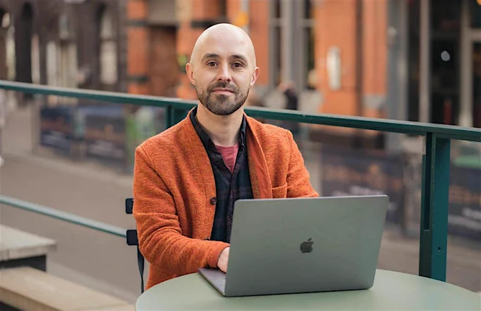 A man sits at a cafe table with a laptop and looking confidently to camera. He wears a bright orange jackets and tartan shirt underneath as the orange city buildings are in the distance behind him.