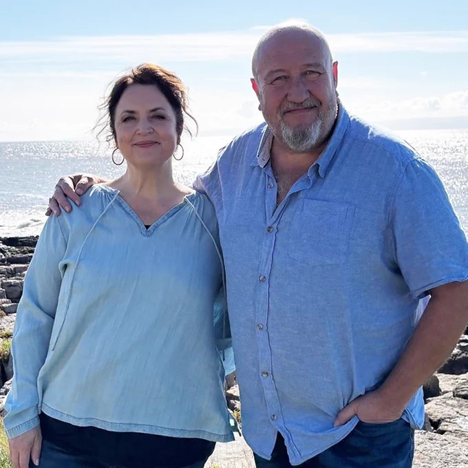 Ruth Jones and Steve Speirs both wearing blue shirts, stood with Steve's arm around Ruth's shoulder, smiling at the camera. They are next to the sea on a sunny day.