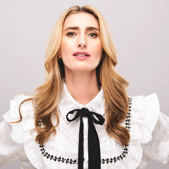 A woman with long blonde hair stands against a grey background in a white shirt with a black bow around the collar