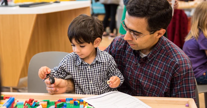 An adult and a child engaging with building blocks at a table in a busy activity room.