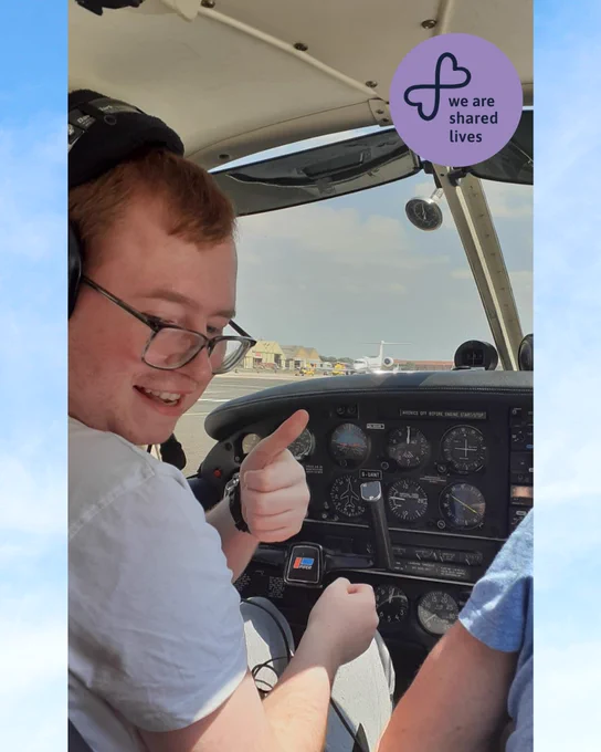 A young man sits in the cockpit of a small plane, smiling and giving a thumbs up. The view through the windscreen shows an airport runway with another aircraft in the distance. A circular logo on a purple background says “We are Shared Lives.”