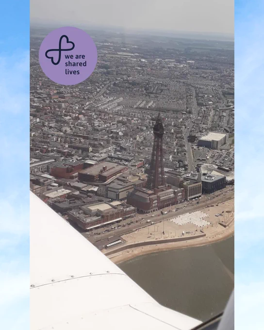 A view from an aircraft window showing Blackpool Tower and the seafront below. The wing of the plane is visible in the foreground, with the town and coastline stretching into the distance. A purple circular logo in the corner reads “We are Shared Lives.”