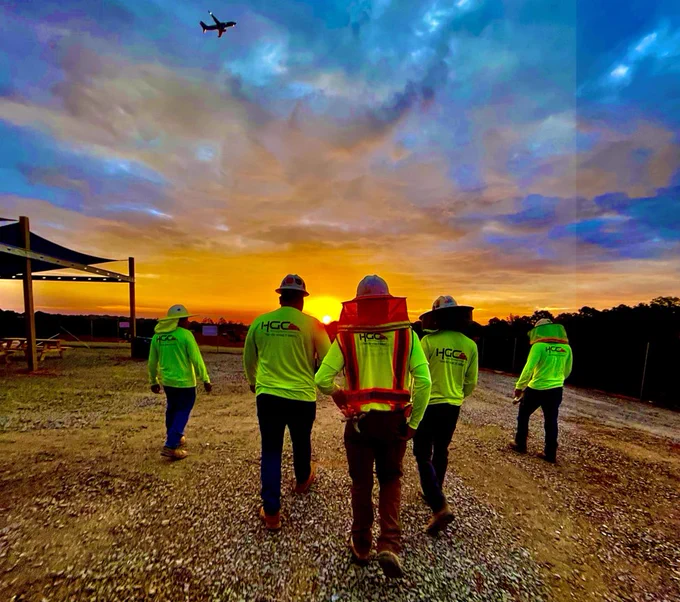 Construction workers walking toward a vibrant orange sunrise on a construction jobsite near Charlotte Douglas International Airport, with a plane flying overhead and the sky glowing in shades of blue and gold.