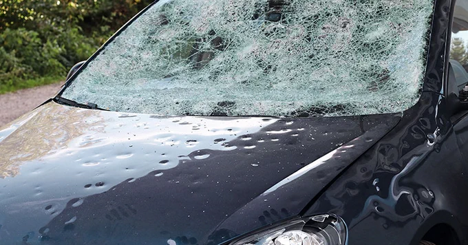 Close-up view of a car with a severely cracked windshield and front damage, parked on a roadside.