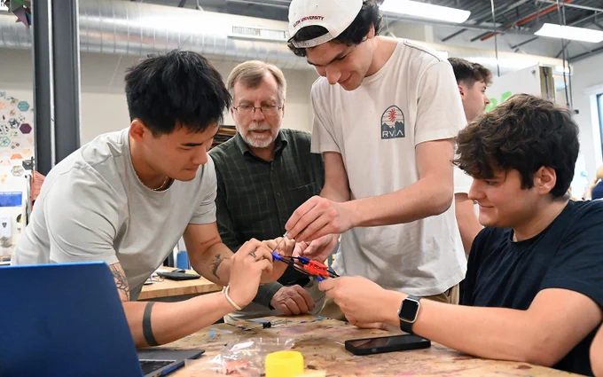 Three individuals and a supervisor observing are engaged in assembling electronic components on a table in a workshop setting.