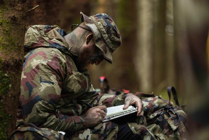 A soldier in camouflage uniform sits against a tree. He is making notes in his notebook.