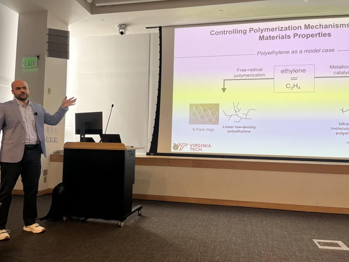 Prof. Adrian Figg from Virginia Tech stands in front of a podium and discusses a slide about controlling polymerization mechanisms from his presentation