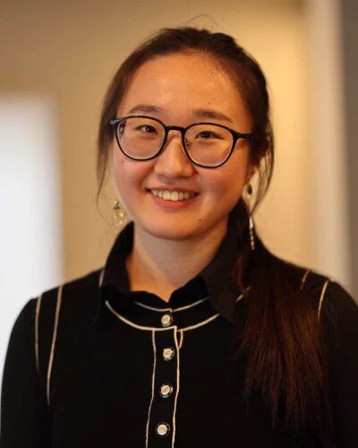 Person with glasses smiling at the camera, wearing a black shirt with white buttons and collar.