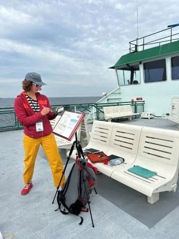 A woman in a red jacket painting on the outer deck of a ferry.