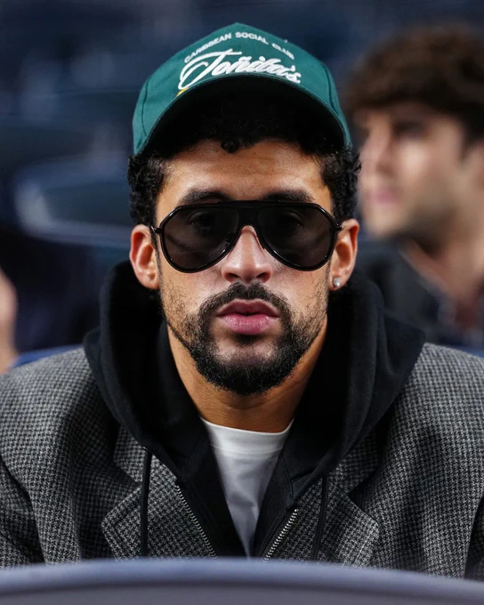 Bad Bunny sitting in the stands at Yankee Stadium. He's wearing a white shirt, gray jacket and green hat.
