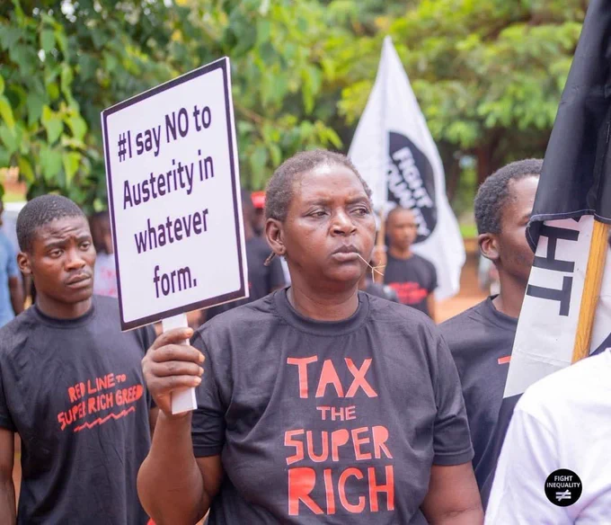 A women in Zambia in a black t-shirt with a slogan saying "Tax the Super Rich" standing among other protesters holds a placard reading "I say NO to Austerity in whatever form"