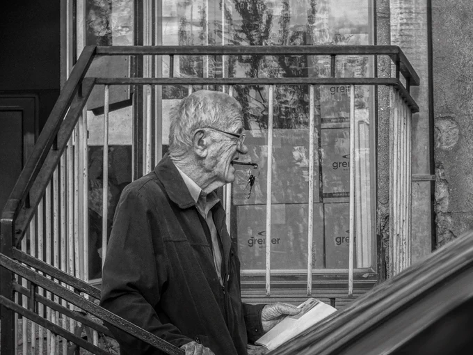 A black and white street photograph by Ivan Maksimović, a documentary photographer from Kosovo and Metohija, capturing an elderly man holding car keys in his mouth, documents in one hand, and steadying himself on a metal railing. The scene conveys quiet resilience, human fragility, and the dignity of everyday life in a raw, realistic setting.