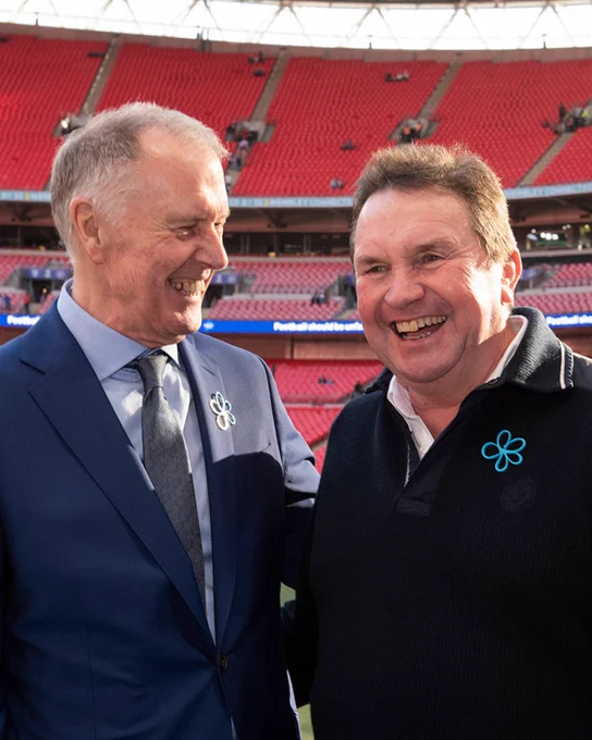 Sir Geoff Hurst and Steve, a white middle aged man, laughing with each other. They are stood pitchside at Wembley Stadium, which is visible in the background. 