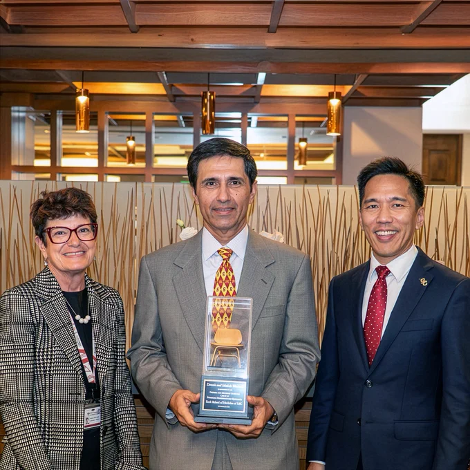 Three individuals stand together in a professional setting, with one in the middle holding an award. They are smiling and positioned indoors with a wooden background. The setting suggests a formal event or recognition ceremony.