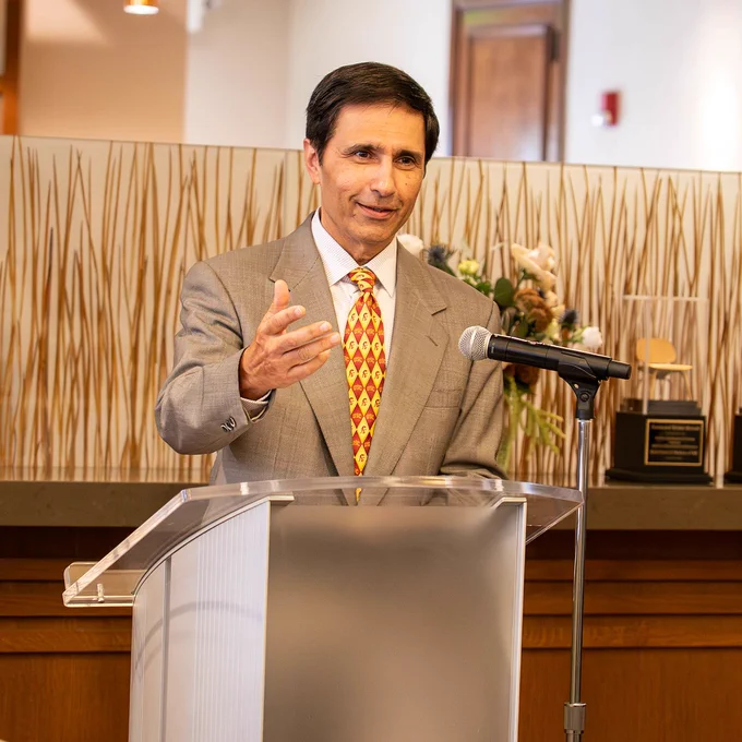 Mark Humayun in a suit standing at a podium with a microphone, gesturing with one hand, in a room with a decorative background.