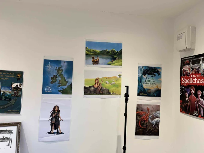 corner of a white room with A3 printouts of artwork stuck on the walls, including the cover of a book reading Celtic Folk and Fairy Tales, a map of the British Isles and Brittany, and illustrations of dragons, a selkie, a water monster, and a herd of cows. Also visible, a black tripod for a camera.