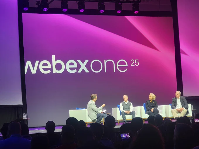 A panel at WebexOne 25 features a seated host interviewing three panelists in front of an audience. The backdrop is a large screen displaying the WebexOne 25 logo in white against a purple background.