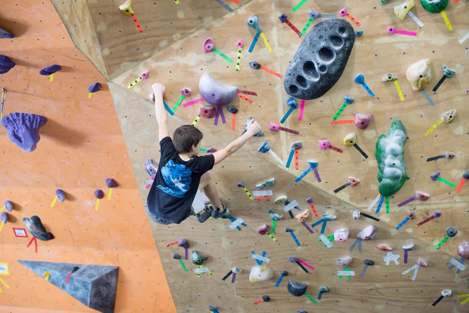 Young boy in a black t-shirt climbing an indoor rock wall with colorful holds and marked routes on a wooden surface.