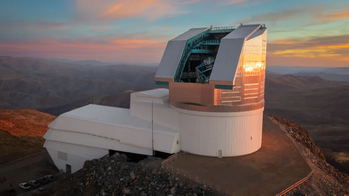Large boot-shaped observatory with a massive telescope dome reflecting orange and golden sunset light atop its rocky mountain site.