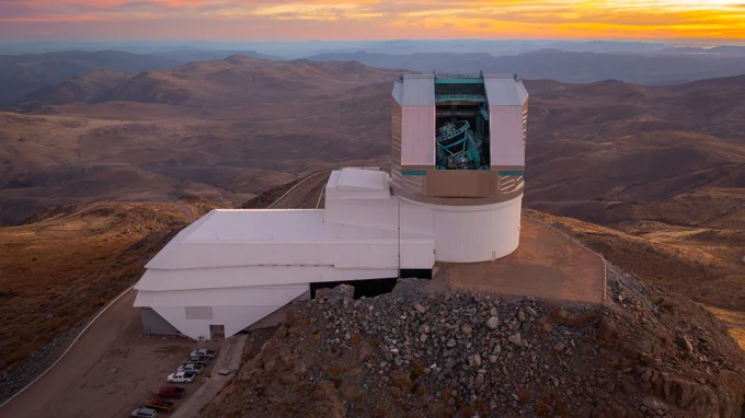 Large boot-shaped observatory with a massive telescope dome atop its rocky mountain site, with the golden glow of sunset over the rolling peaks in the distance.