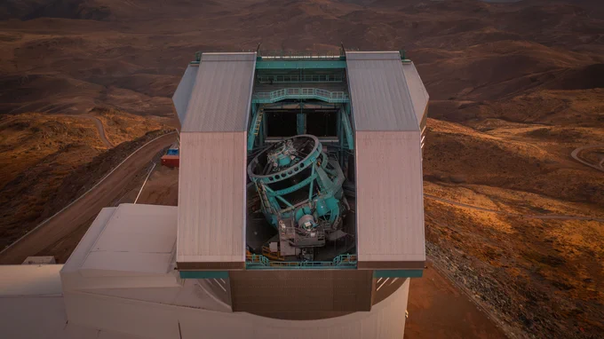 Drone shot of a massive teal telescope within an observatory's dome with a vertical opening, surrounded by winding mountain roads and dry terrain.