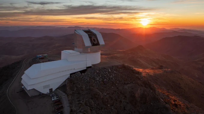 Large boot-shaped observatory on a rocky mountain peak during a vibrant sunset with layered mountain ranges in the background.