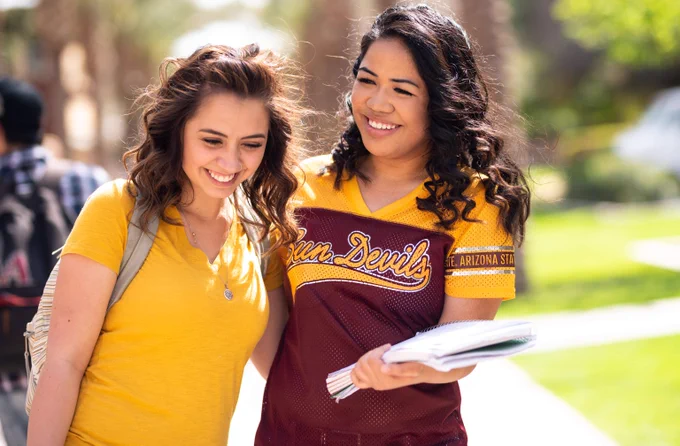 Two students standing outdoors on a sunny day, one wearing a yellow shirt and the other in an Arizona State Sun Devils jersey holding notebooks.