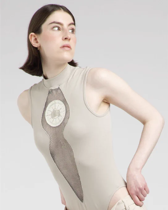 Close-up of model wearing beige sleeveless top with mesh cut-out and circular embroidered patch on the chest.