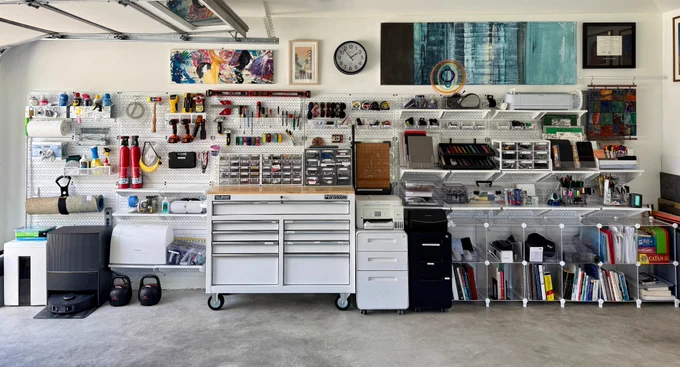 A neatly organized garage workspace featuring Wall Control metal pegboards mounted on the wall, holding tools, supplies, and craft materials. Below are a large rolling tool chest, filing cabinets, printers, and modular shelving filled with books, boxes, and office items. Artwork and a clock hang above the pegboards, creating a clean, efficient, and creative environment.