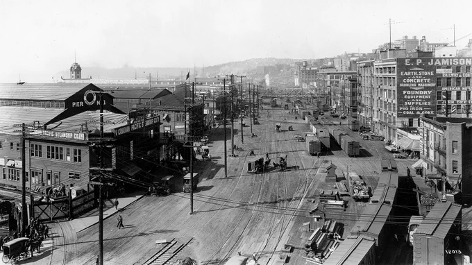A historic black and white photo of Railroad Avenue on the Seattle waterfront. Nine lanes of train tracks and a series of piers are seen.