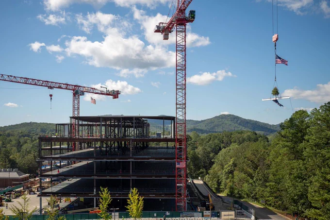 Construction site with cranes lifting materials, working on a large building structure, set against a backdrop of green hills under a clear sky.
