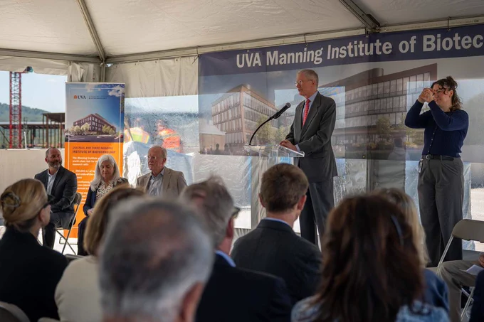 A speaker addresses an audience at a UVA Manning Institute of Biotechnology event. Banners with institute details and architectural images are visible behind the speaker, while attendees listen attentively.