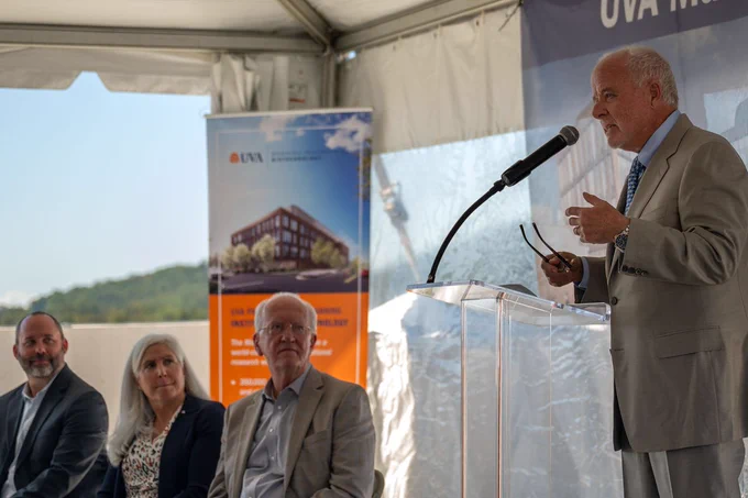 An individual speaking at a podium at a UVA event, with three attendees seated nearby under a tent, listening attentively.