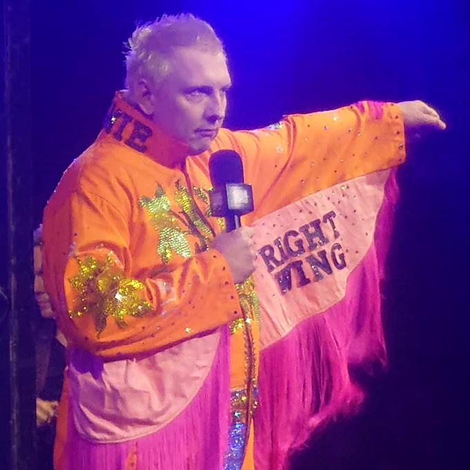 Joe Lycett onstage, lifting up his left arm to reveal RIGHT WING written above the pink fringing